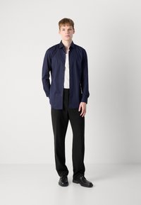Navy button-up shirt with long sleeves, paired with black trousers and black shoes, set against a plain gray background.