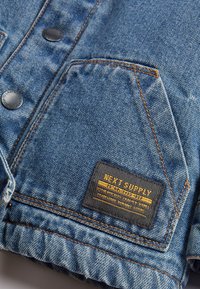 Veste en denim au design classique, avec des boutons en métal foncé, des coutures orange et une étiquette noire avec du texte jaune sur la poche de poitrine gauche.