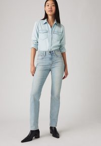 Šviesiai mėlyna denimo marškiniai su dviem kišenėmis, dėvimi su aukštu juosmeniu šviesiai mėlynomis džinsomis ir juodais aukštakulniais auliniais batais. Lygus audinio tekstūra, kasdienis siluetas.