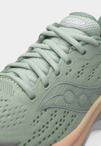 Ανοιχτό πράσινο αθλητικό παπούτσι Saucony με αναπνεύσιμο πλέγμα, κορδόνια και ανοιχτό καφέ σόλα σε κοντινή λήψη.