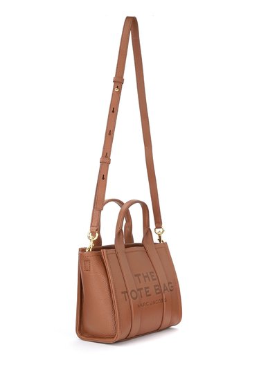 Brązowa skórzana torba tote z wytłoczonym tekstem "THE TOTE BAG" i "MARC JACOBS." Posiada podwójne uchwyty, regulowany pasek na ramię oraz wyraźny kształt.
