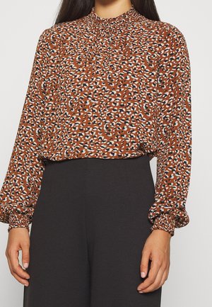 Blusa marrone a fantasia con maniche lunghe a sbuffo, scollo arricciato e macchie blu e bianche sparse, abbinata a pantaloni scuri.