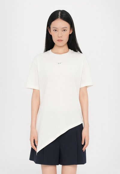 T-shirt blanc asymétrique à manches courtes, avec une petite touche métallique sur la poitrine, associé à une jupe bleu marine foncé.