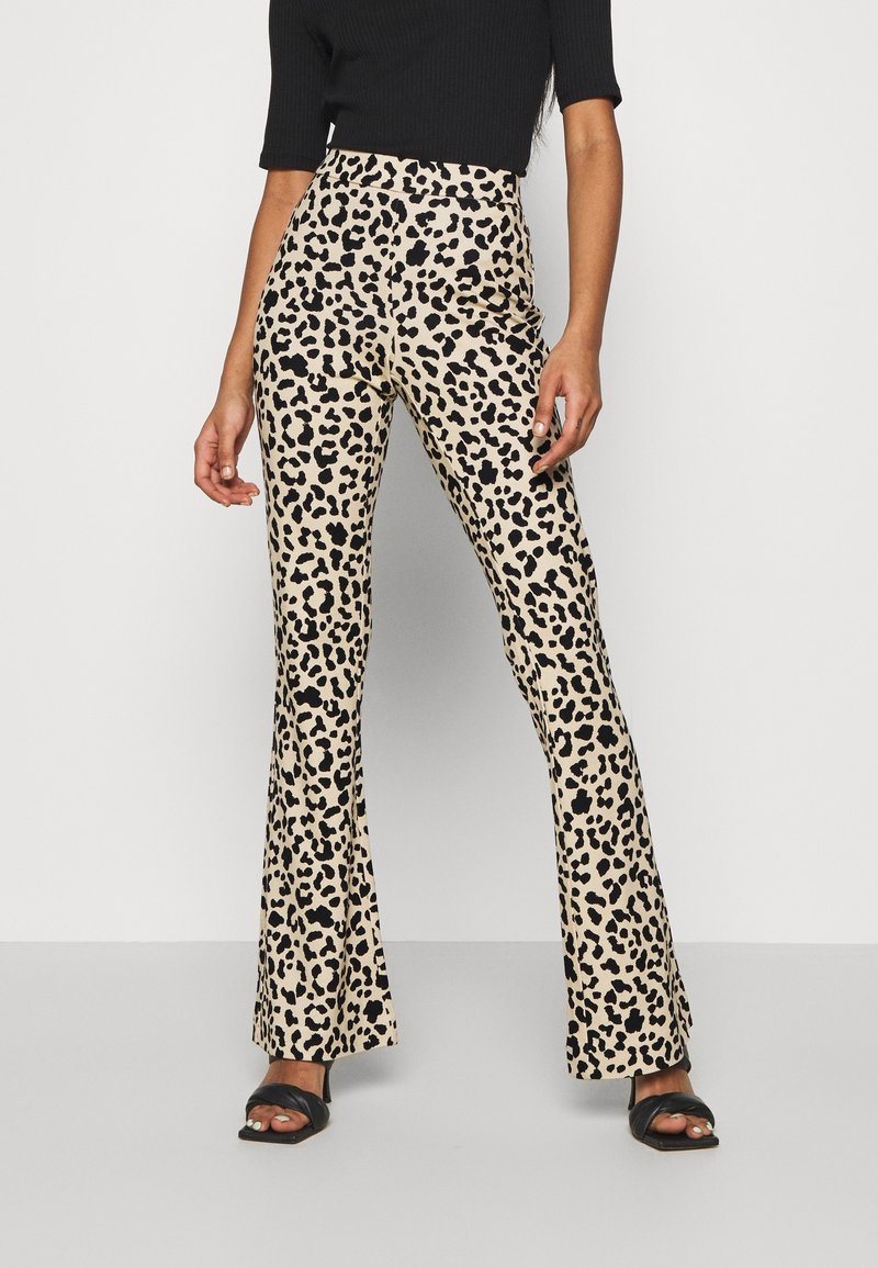 Femme portant un pantalon évasé à imprimé léopard beige et noir, un haut noir à manches courtes et des sandales noires à bouts ouverts, debout devant un fond uni.
