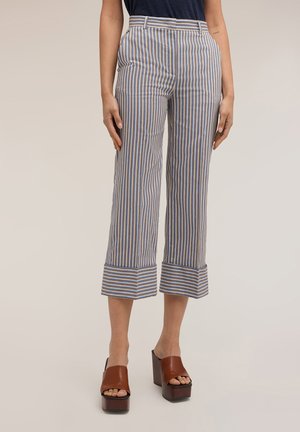 Kobieta nosząca niebieskie, białe i brązowe pionowo paskowane krótkie spodnie z mankietami oraz brązowe sandały na platformie, stojąca na jednolitym tle.