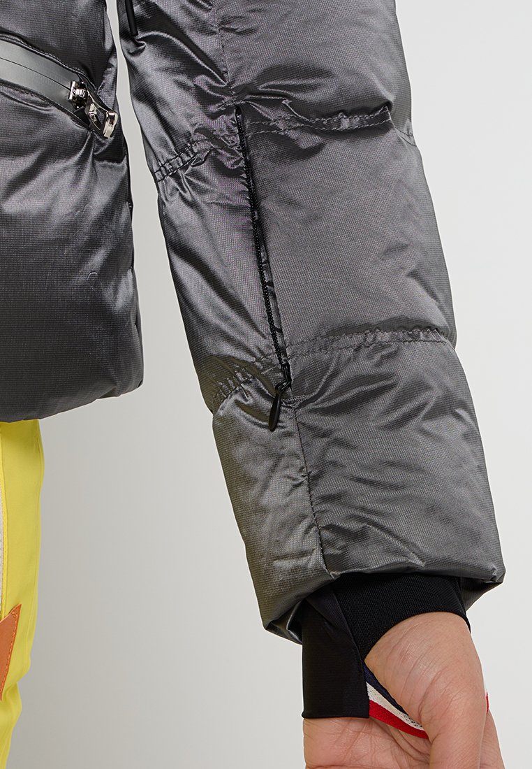 Chaqueta acolchada gris con una textura brillante, que presenta un bolsillo con cremallera, puños elásticos y un forro interior negro. Ajuste con cordón en la manga.