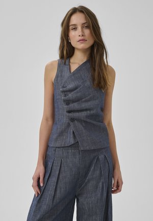 Žena oblečená v tmavosivej asymetrickej veste bez rukávov s gombíkmi a zodpovedajúcich plisovaných širokých nohaviciach, stojaca pred jednoliatym pozadím.