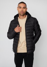 Zwarte pufferjack met ritssluiting, geweven ontwerp en capuchon. Draag het over een lichtbeige sweatshirt en donkere jeans. Soepele textuur.