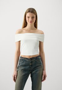White off-the-shoulder crop top with draped fabric. Paired with high-waisted blue denim jeans featuring a faded wash and decorative buttons.