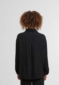 Black long-sleeve shirt with white vertical stripes, featuring a collar and a pleat at the back, made from a soft textile.