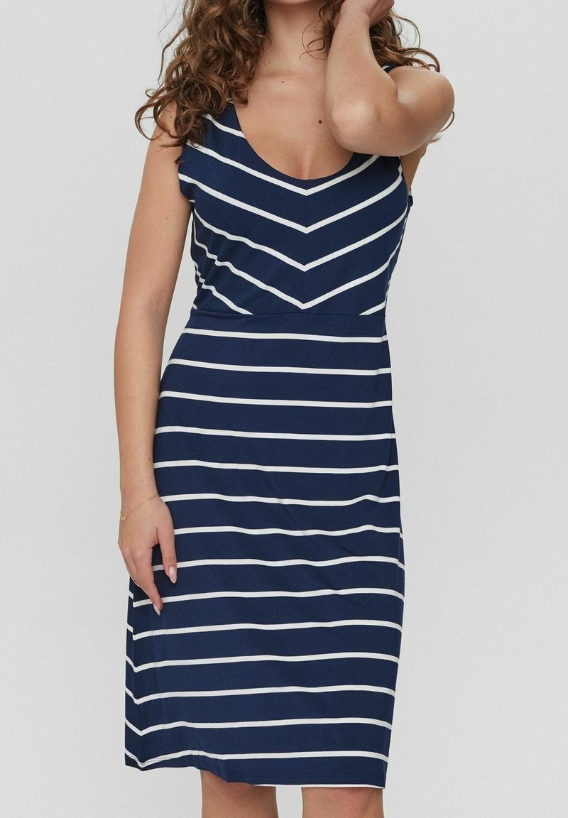 Femme portant une robe bleu marine sans manches avec des rayures blanches horizontales et un motif en forme de V sur la poitrine sur fond clair.