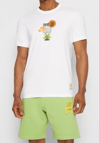 Camiseta blanca de algodón con un gráfico colorido que presenta un girasol, hojas y el logo de Nike. Combinada con pantalones cortos de algodón verdes con bordado.