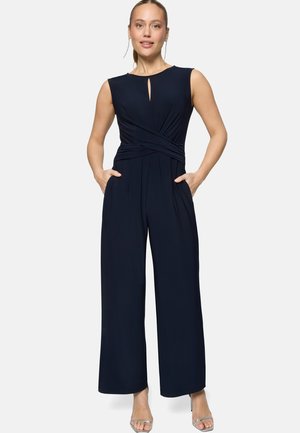 Frau trägt ärmellosen marineblauen Overall mit Schlitz am Dekolleté und weitem Bein, Hände in den Taschen, steht vor weißem Hintergrund.