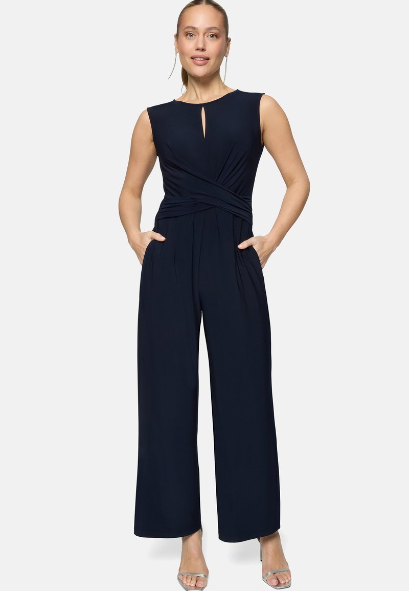 Femme portant une combinaison sans manches bleu marine avec encolure découpée en clé et pantalons à jambes larges, mains dans les poches, debout devant un fond blanc.