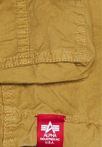 Khaki-stoff med detaljerte sømmer og et rødt Alpha Industries Inc. U.S.A. logo merke sydd på sømmen.