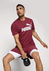 Marron sportstrøje med hvid "PUMA" logo, parret med marron shorts med hvide accenter. Personen holder en sort kettlebell.