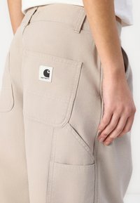 Pantaloni cargo beige chiaro realizzati in tessuto resistente, con grandi tasche laterali e una toppa quadrata con logo sulla tasca posteriore.