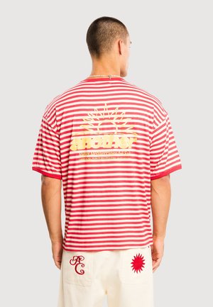 Uomo che indossa una maglietta a righe rosse e bianche con grafica di sole giallo e testo sul retro, abbinata a pantaloni bianchi con toppe ricamate rosse.