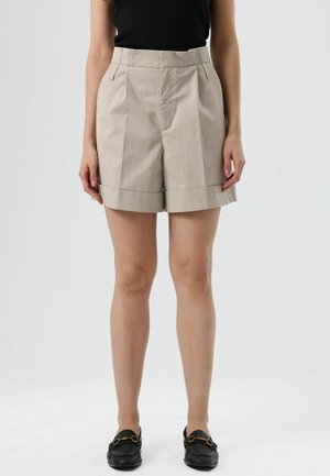 Shorts beige ajustés avec des plis, ourlet retroussé et poches latérales, fabriqués dans un tissu lisse. Portés avec un haut noir et des mocassins noirs.