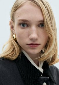 Close-up of a person with shoulder-length, straight blonde hair, wearing a black cord jacket and large gold earrings. Smooth skin, light blue eyes.