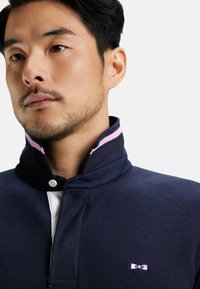 Pull-over marine à col montant, en tissu texturé, avec des accents roses sur le col et un petit logo en forme de nœud sur la poitrine. Fermeture à boutons au niveau du col.