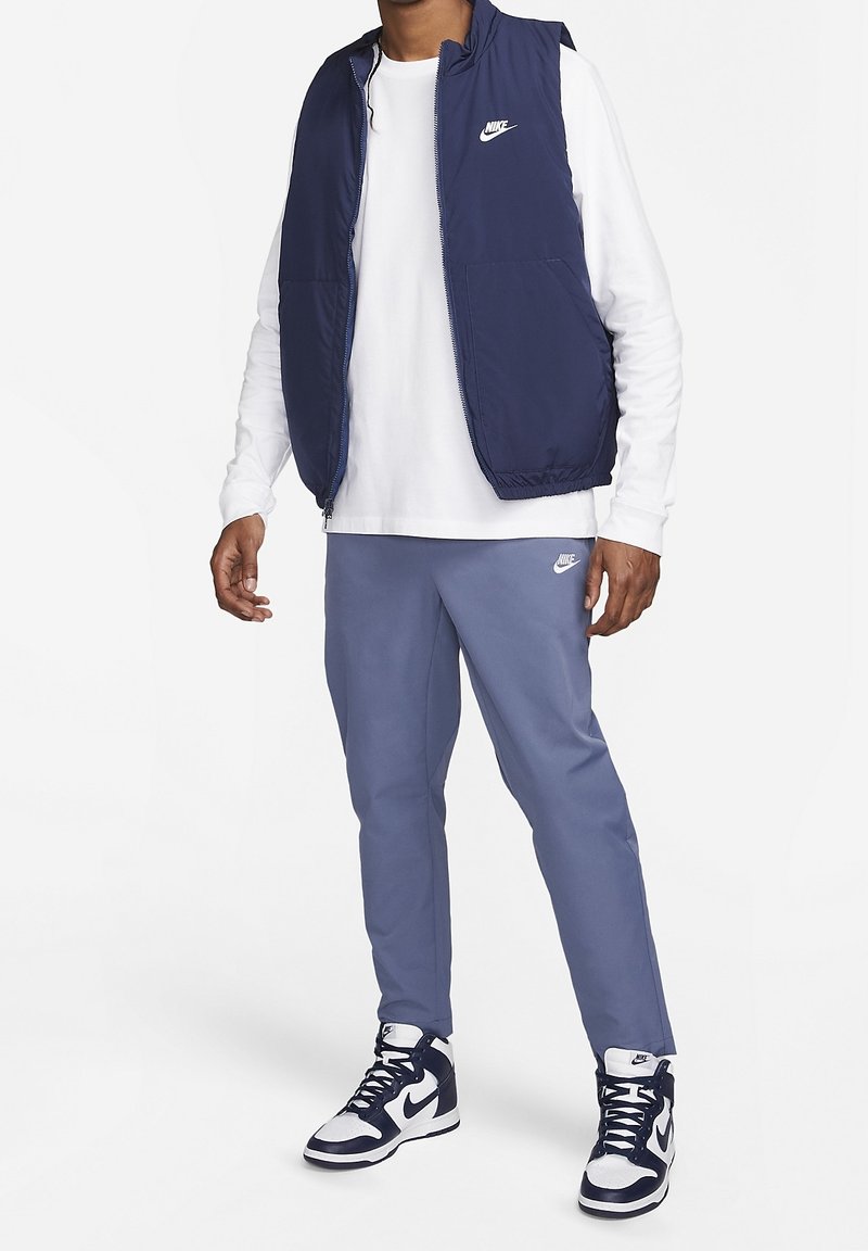 Tummansininen hihaton liivi valkoisen pitkähihaisen paidan päällä, harmaat verkkarit jalassa ja korkeavartiset tennarit, joissa on mustavalkoisia yksityiskohtia.