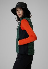 Jeune femme portant un chapeau bob noir, un gilet sans manches matelassé vert et noir, et un haut à manches longues orange, debout devant un fond gris.