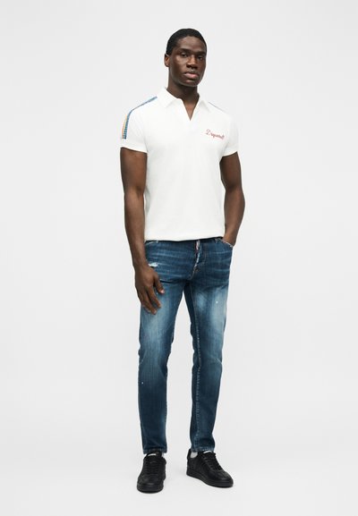 Homme debout les mains dans les poches, portant un polo blanc avec des détails rayés sur les épaules, un jean bleu et des baskets noires, sur un fond uni.