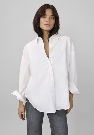 Žena s tmavými vlasmi po plecia, oblečená v voľnej bielej košeli na gombíky a šedých džínsoch, stojaca pred jednoduchým sivým pozadím.