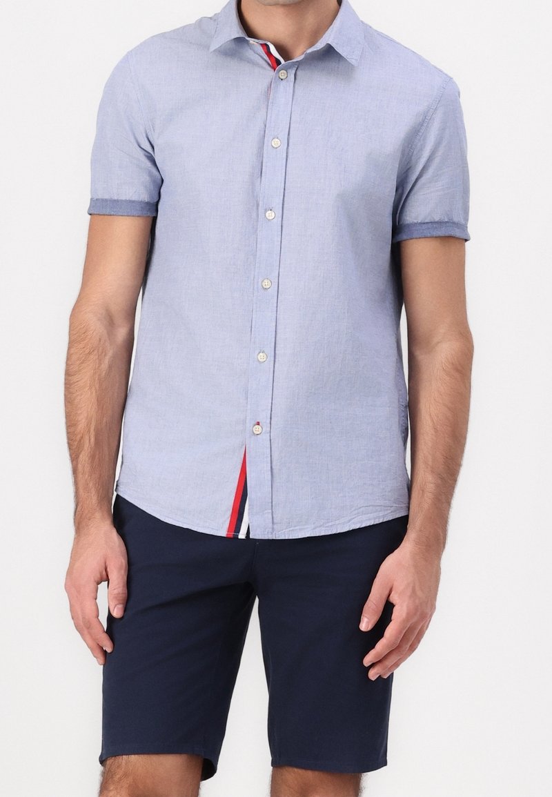 Camicetta azzurra chiara a maniche corte con tessuto texturizzato, caratterizzata da un accento a righe in rosso e blu navy all'orlo. Abbinata a pantaloni corti blu navy.