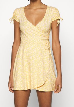 Vrouw draagt een korte gele wikkeljurk met witte bloemenprint, korte geplooide mouwen en strikdetails aan taille en mouwen.