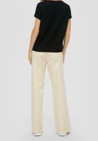 Žena v černém krátkém rukávu a krémových širokých kalhotách s vysokým pasem, stojící zády k nám na jednobarevném pozadí.