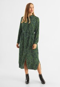 Rochie verde cu model în valuri, mâneci lungi, închidere cu nasturi în față și curea la talie; are fante laterale. Pur tată cu botine negre până la glezne.