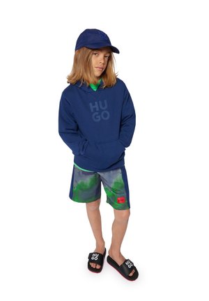 Haut à capuche bleu marine avec le logo "HUGO", t-shirt vert en dessous, shorts à motifs et sandales noires avec des accents rouges. Look décontracté avec une coupe ample.