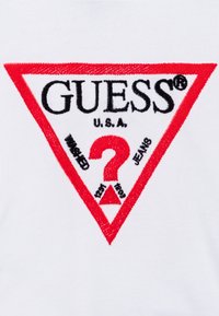 Fehér anyag, amelyen piros háromszög alakú hímzett logó látható "GUESS", "U.S.A." és egy kérdőjel felirattal, valamint "WASHED" és "JEANS" fekete színben.