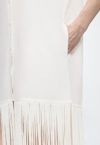 Χέρι μέσα στην τσέπη ενός λευκού φορέματος με κάθετες φράγες στο κάτω μέρος.