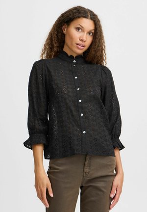 Schwarze Bluse mit einem strukturierten Blumenmuster, hohem Kragen und Knopfschluss. Sie hat Dreiviertelärmel mit Rüsche.