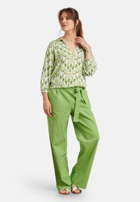 Grün gemusterte Bluse mit weiten Ärmeln, kombiniert mit hoch taillierten, hellgrünen Hosen mit einem Bindegürtel. Glatte Stofftextur.