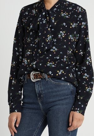 Zwarte blouse met lange mouwen, kleine meerkleurige bloemenprint en een gestrikte strik bij de hals, gecombineerd met een blauwe spijkerbroek en een luipaardprint riem.