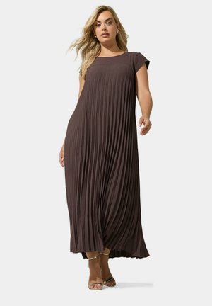 Blonde vrouw die loopt in een vloerlang donkerbruin geplooid jurkje met korte mouwen en gouden enkelbandjes op een witte achtergrond.