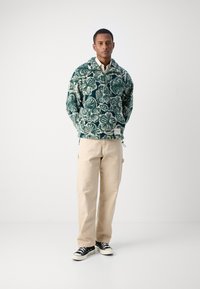 Pullover verde con motivo floreale realizzato in tessuto testurizzato, abbinato a pantaloni beige a gamba dritta e sneakers nere con suola bianca.