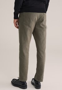 Hombre vestido con pantalones rectos de color verde oliva, zapatos de vestir negros y un suéter negro, de pie frente a un fondo beige liso.