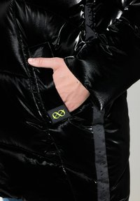 Svart puffjacka med glänsande yta, med en sidoficka och en neongrön logotags. Detaljer inkluderar stickade mönster och kontrasterande kant.