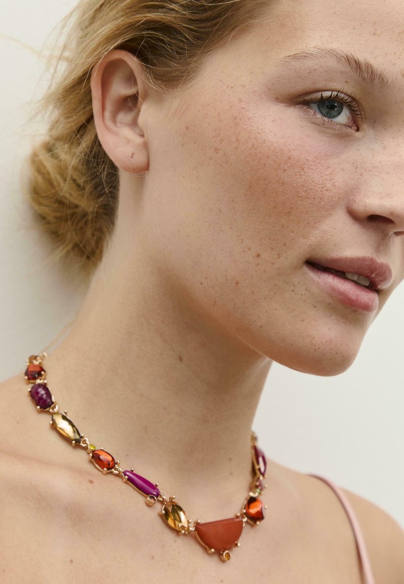 Collana con pietre in vetro sfaccettato di diversi colori nelle tonalità di viola, arancione e oro, montate su una catena di metallo in oro rosa.