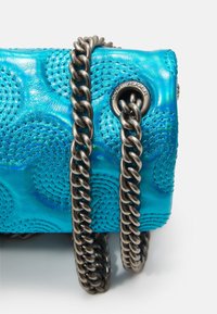 Angolo di una borsa in pelle trapuntata blu metallico con una spessa tracolla di metallo scuro, infilata attraverso un occhiello metallico inciso "Kurt Geiger."