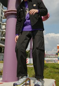 Persona che indossa pantaloni cargo neri, un maglione viola, una giacca nera e sneaker grigie, in piedi su un pavimento di cemento con tubi viola e graffiti sullo sfondo.