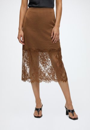 Jupe midi marron avec ourlet en dentelle superposée, portée avec un top noir sans manches et des sandales noires à talons ouvertes, debout sur un fond clair.