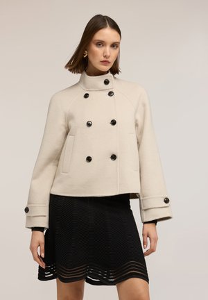 Donna che indossa una giacca doppiopetto color crema in lana con bottoni neri e una gonna nera testurizzata, in piedi davanti a uno sfondo beige semplice.