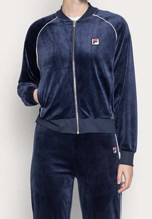 Osoba ubrana w granatowy welurowy dres z białym pipingiem, zapinany na zamek kurtka, ściągacze na rękawach oraz małe logo w kolorach czerwonym, białym i granatowym na piersi i spodniach.