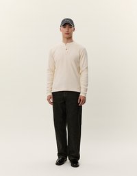 Beige hosszú ujjú henley ing, három gombbal, fekete, egyenes szárú farmernadrággal párosítva. A modellen szürke sapka és fekete cipő van.